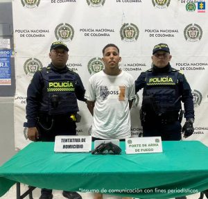 En la foto está un hombre joven, moreno, cabellos crespos negros, vestido con una camiseta blanca, tiene una semi barba y se encuentra custodiado por dos uniformados de la Policía Nacional. Delante de ellos hay una mesa con un arma de fuego.