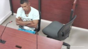 En la imagen está un hombre trigueño sentado en una silla al frente de un escritorio de madera con un microfono al interior de la sala de audiencia donde se le procesó.