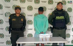 En la imagen está un hombre delgado de pie, de espalda con los brazos esposados, vestido con un buzo verde aguamarina y una pantaloneta azul. Se encuentra custodiado por dos agentes de la Policía Nacional. Delante de ellos hay una mesa donde están las municiones incautadas.