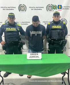 En la imagen hay un hombre joven, trigueño, de pie, de frente, cabellos negros, tiene la cabeza agachada, está vestido con buzo negro y jean azul. Se encuentra acompañado por dos policías.