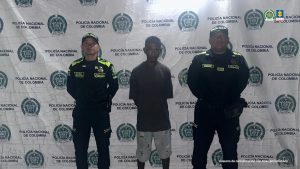 En la imagen está un hombre delgado de pie, de frente, moreno, de baja estatura, vestido con camiseta negra y bermuda blanca, acompañado por dos policías. 