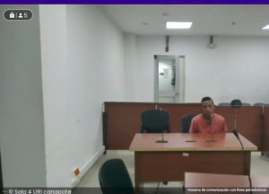 En la fotografía esta un hombre delgado, sentado en un escritorio de madera con micrófono, vestido con camiseta naranja, en la sala de audiencia donde se le procesó.