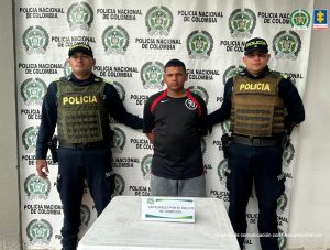  En la imagen está un hombre joven moreno, de pie, de frente, con los brazos atrás esposados, de contextura gruesa, cabellos negros crespos, nariz y labios grandes. Está vestido con una camiseta negra y un pantalón gris y a su lado están dos policías. 