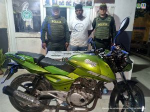 En la imagen aparece un hombre de contextura mediana, de tez morena, vestido de camiseta blanca con jean, custodiado por dos agentes de la Policía Nacional. Detrás de ellos aparece un pendón de esa institución y delante de ellos una motocicleta verde que fue la que presuntamente hurtó el procesado.