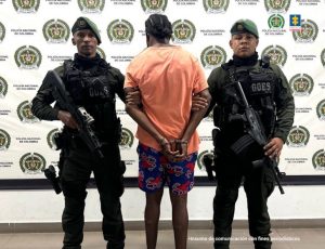 En la imagen está un hombre moreno alto, delgado, de pie, de espalda con los brazos esposados, vestido con camiseta naranja, bermuda estampada azul y tenis blancos. Se encuentra custodiado por dos uniformados de la Policía Nacional.