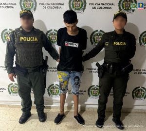 En la imagen está un hombre delgado, joven, trigueño de cabellos negros, con la cabeza agachada, de pie, con los brazos atrás, vestido con camiseta negra, bermuda de jean y tenis negros, custodiado por dos uniformados de la Policía Nacional.