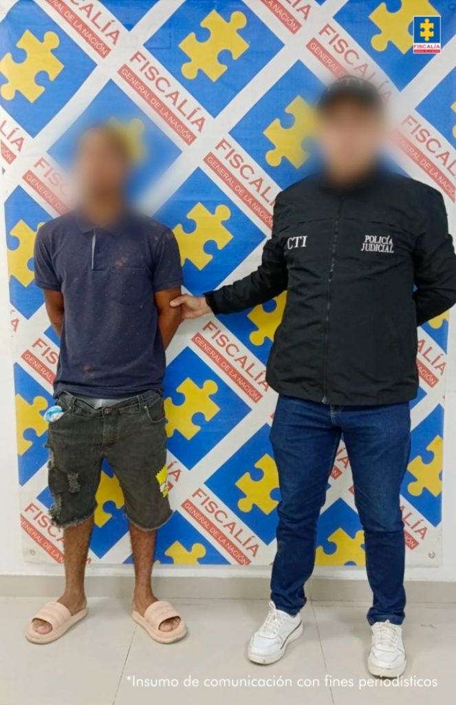 En la imagen se encuentra un hombre de pie, de baja estatura con el rostro difuminado, con los brazos atrás, vestido con camiseta azul oscuro, bermuda gris y sandalias rosadas, custodiado por un uniformado del Cuerpo Técnico de Investigación (CTI).