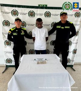 En la imagen está un hombre moreno, delgado de pie de frente con los brazos atrás esposados, cabellos crespos negros, cabeza agachada, vestido con camiseta blanca y pantalón negro. Se encuentra custodiado por dos uniformados de la Policía Nacional.