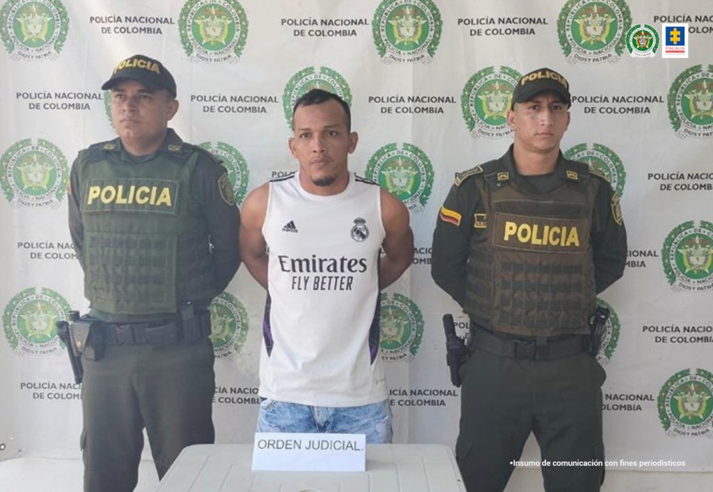 En la fotografía hay a un hombre de camiseta blanca, custodiado por dos funcionarios de la Policía Nacional. Ellos están ubicados delante de un pendón de esa institución.