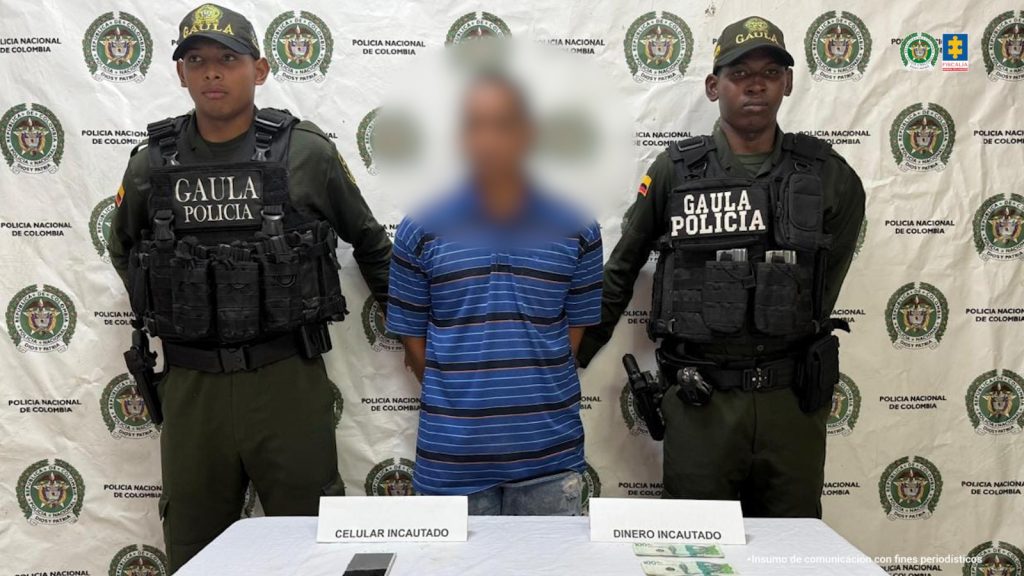 En la imagen está un hombre de pie, con el rostro difuso, de baja estatura, moreno, vestido con una camiseta azul y rayas negras, tiene los brazos atrás esposados y se encuentra acompañado por dos uniformados de la Policía Nacional.