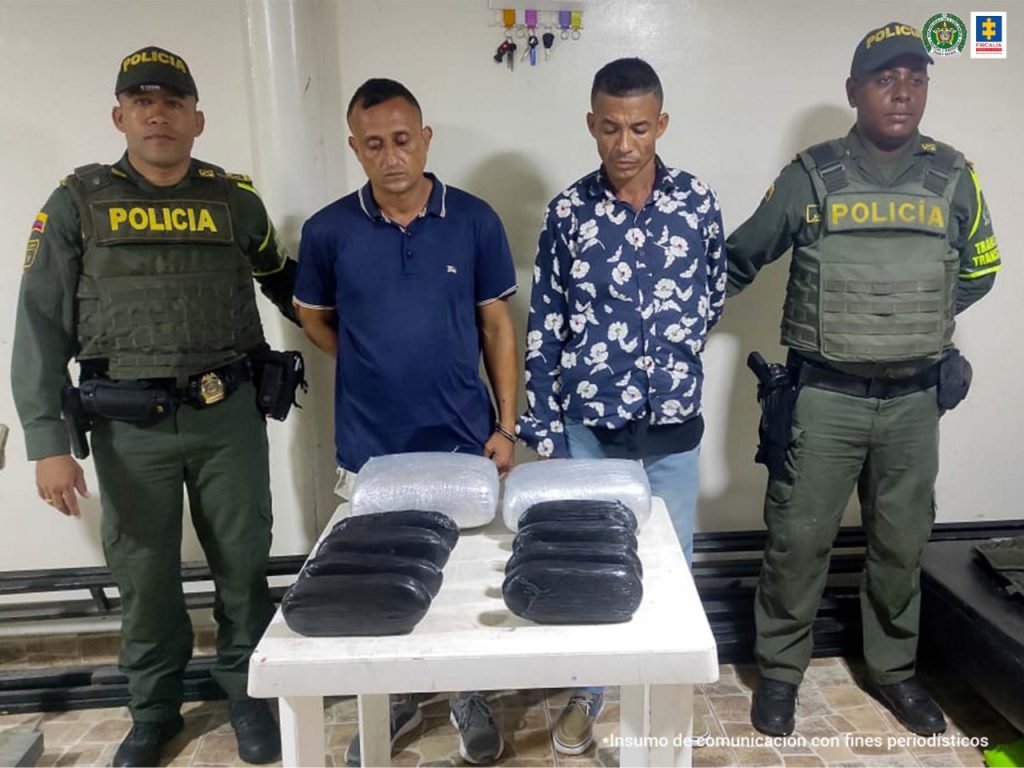 En la fotografía se observan a dos hombres, uno de camiseta azul manga corta y el otro de camisa azul manga larga, custodiado por dos agentes de la Policía Nacional. En una mesa se observa una mesa con unos paquetes.