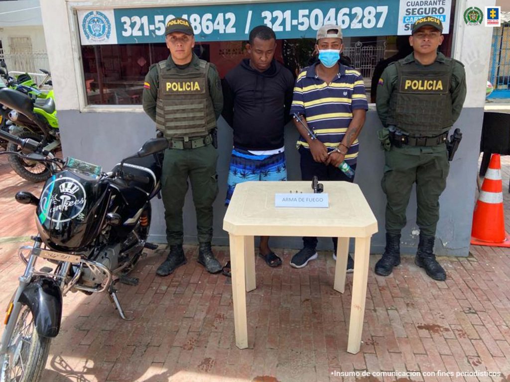 En la fotografía se observan dos hombres uno con camiseta de rallas y el otro con chaqueta oscura custodiado por dos agentes de la Policía Nacional en un CAI de la Policía Nacional.