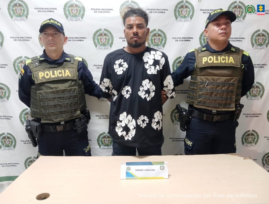 En la imagen esta un hombre de pie, moreno, con cabellos negros ondulados, barba negra, con los brazos atrás esposados, vestido con una camiseta negra con blanco y jean negro. El procesado se encuentra con dos uniformados de la Policía Nacional.