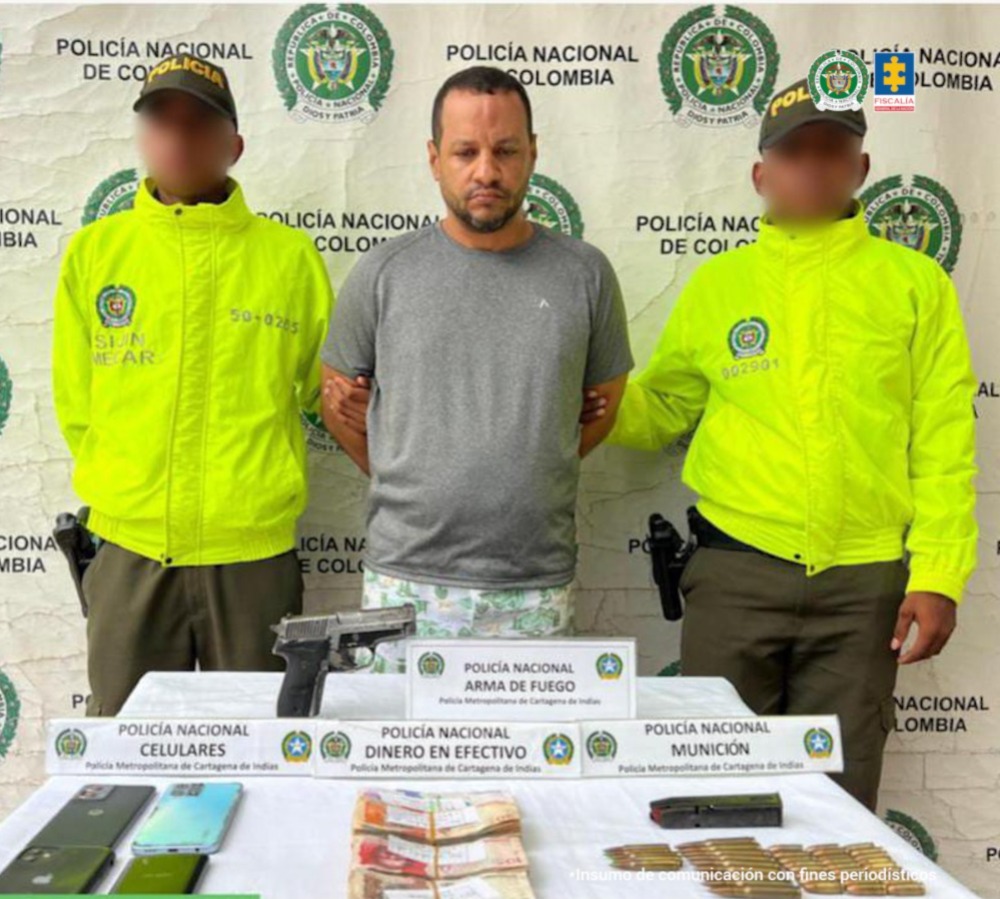 En la imagen está un hombre trigueño, de pie, de cabellos negros cortos y barba negra, se encuentra con los brazos atrás esposado y vestido con camiseta gris y pantalón blanco. A su lado están uniformados de la Policía Nacional.