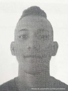 En la fotografía a blanco y negro, aparece la cara de un hombre delgado, joven, cara ovalada, de nariz, boca y orejas medianas, ojos grandes, tiene un corte de cabello con cresta y se encuentra vestido con una camiseta sin cuello.