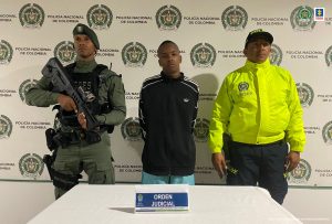  En la fotografía aparece un hombre moreno vestido con buzo negro y pantalón azul claro, custodiado por dos uniformados de la Policía Nacional delante de un pendón de esa institución.