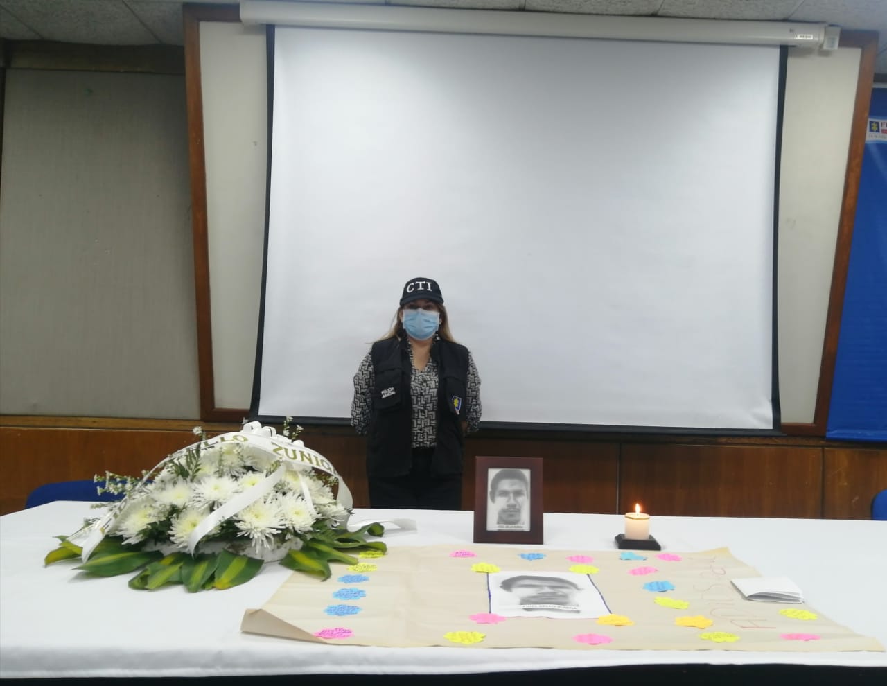 Se observa a una uniformada del CTI detrás de una mesa en la que se encuentra un arreglo floral y una cartelera con la foto de la víctima y los mensajes que los familiares les escribieron.