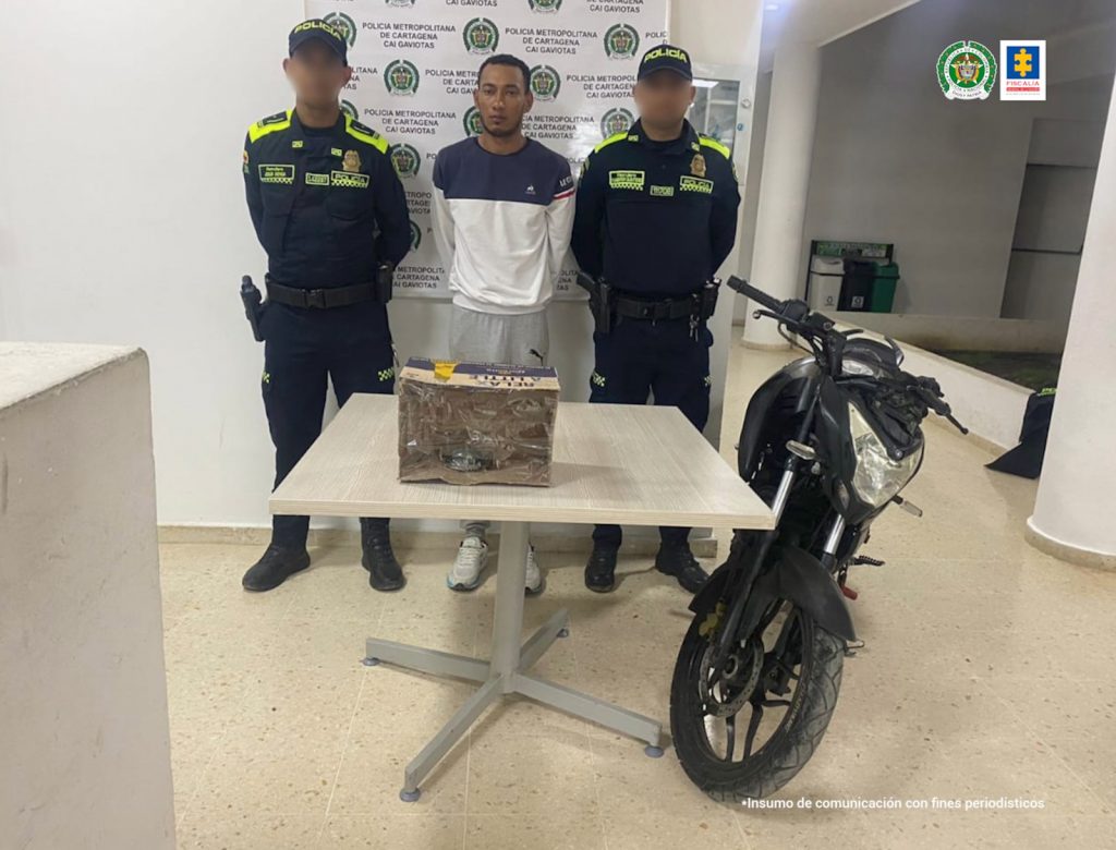 En la imagen esta un hombre de pie, moreno, delgado, vestido con un buzo blanco y azul oscuro, sudadera gris y tenis blanco. Esta acompañado por dos uniformados de la Policía Nacional y delante de ellos está una mesa con la granada y la moto incautada.