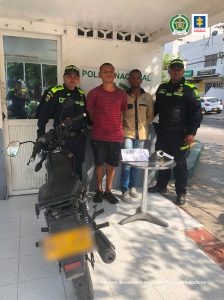 En la fotografía se identifican a dos hombres, uno con camiseta roja, bermuda negra y tennis negro y el otro con camisa amarilla, jean azul y tennis blanco custodiados por dos agentes de la Policía Nacional. También se observa una mesa delante de ellos con el arma que le incautaron en las capturas.