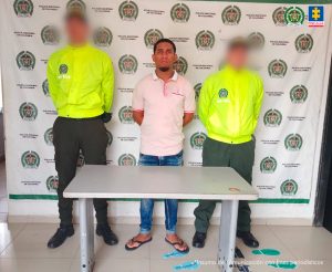 En la imagen está un hombre de mediana estatura, de pie, con los brazos atrás, vestido con camiseta rosada, jean azul y sandalias azul oscuras, custodiado por dos uniformados de la Policía Nacional.