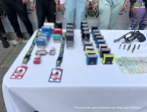 En la fotografía 2 se ve una superficie blanca y sobre ella se parecían sellos, escarapelas, dinero en efectivo, un arma de fuego y munición. En la fotografía 3 se ve una superficie blanca y sobre ella hay una pantalla, un computador portátil, celulares, una USB, dinero en efectivo, munición y un arma de fuego.