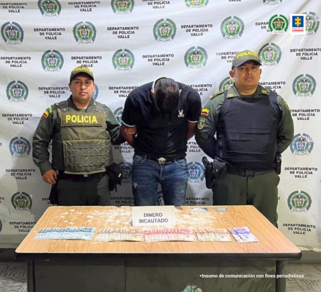 En la foto aparece de pie, los brazos atrás y agachado, Andrés Armando Merchán Gutiérrez. Viste camisa negra de manga corta y jean azul. A lado y lado de él hay uniformados de la Policía Nacional. Delante de ellos hay una mesa y sobre ella hay dinero en efectivo y un letrero que dice. ‘Dinero incautado’. Detrás de ellos hay un pendón institucional de la Policía.