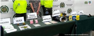 En la foto aparece en primer plano una mesa con mantel verde y sobre ella hay varios letreros que dicen: ‘Cocaína’, ‘WAX’, ‘Droga sintética’, ‘Pastillas’, ‘Marihuana’, ‘Munición’ y ‘Arma de fuego’. Asimismo, se aprecian elementos alusivos a dicha información.