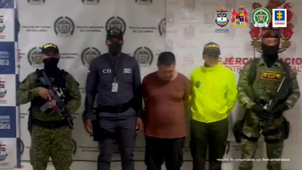 En la foto aparece Jhon Fredy Mejía Torres alias ´Gambeta´, con los brazos atrás y la cabeza agachada; vestido con una camiseta de manga corta café, bermudas oscuras y zapatos oscuros. Al lado y lado de él hay uniformados del CTI de la Fiscalía, Policía, Ejército y Armada Nacional. Detrás de ellos hay tres pendones institucionales del Gaula militar de Buenaventura, Policía Nacional y Ejército.