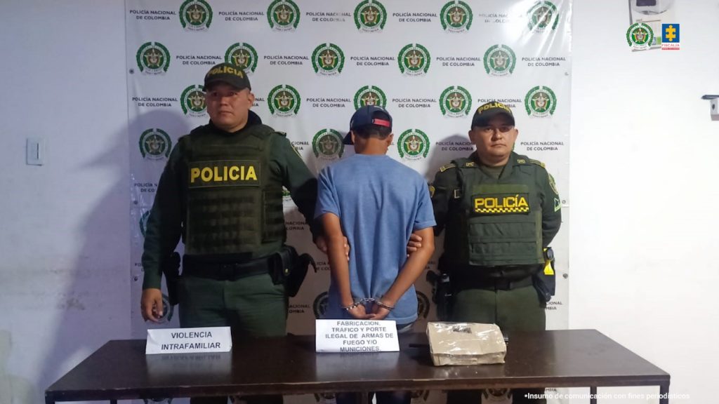 En la fotografía se aprecia de pie, con los brazos atrás, esposado y de espaldas a la cámara a Jhon Stiven Téllez. Viste camiseta de manga corta azul y pantalón oscuro. Usa gorra de color azul oscuro con sistema de graduación en color rojo. A sus extremos hay uniformados de la Policía Nacional. Delante de ellos hay una mesa color marrón y sobre ella hay un paquete y dos letreros que dicen: ‘Violencia intrafamiliar’ y ‘Fabricación, tráfico y porte ilegal de armas de fuego y/o municiones’. Detrás de ellos hay un pendón institucional de la Policía.