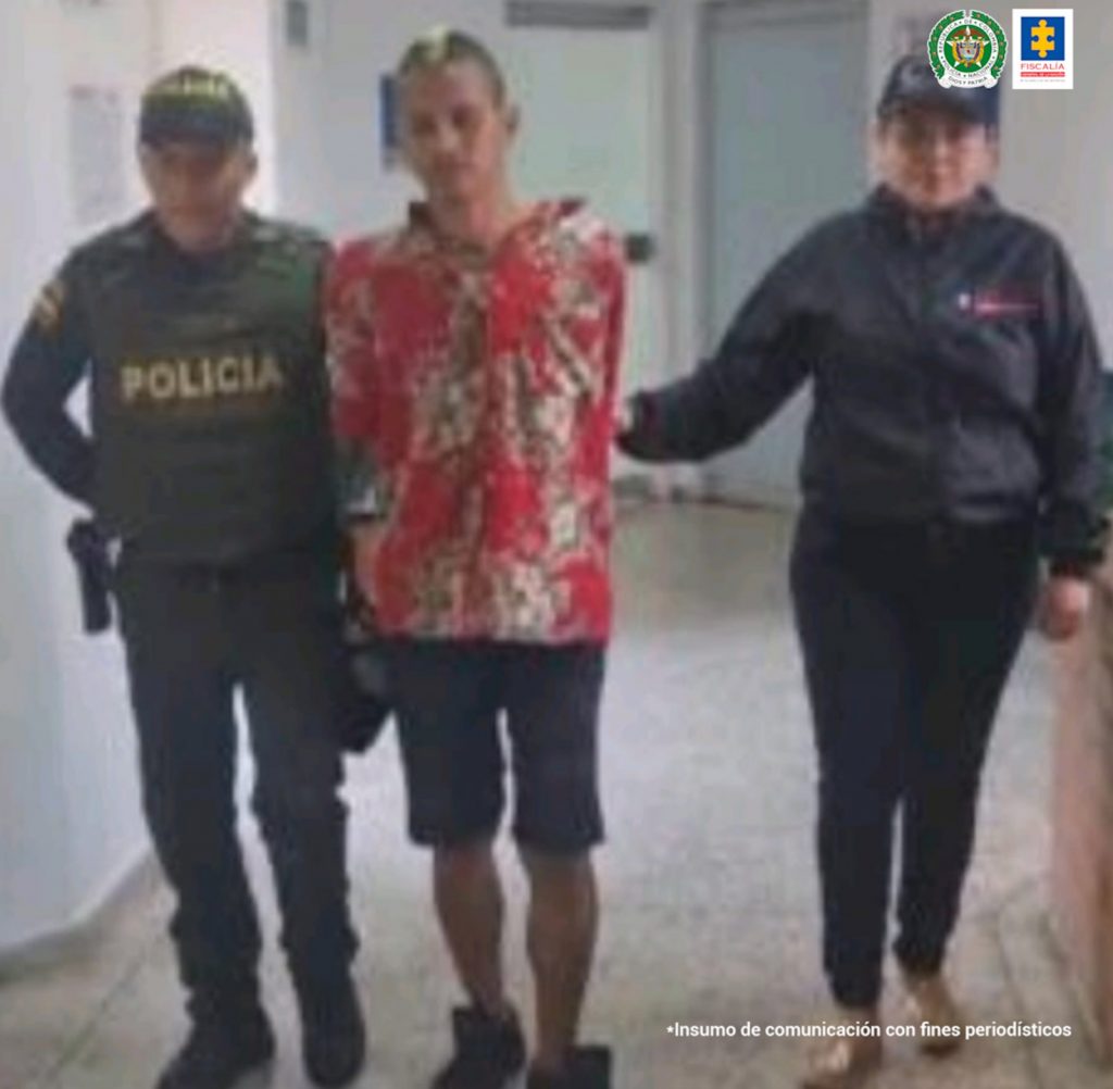 En la fotografía se aprecia en acción de caminata, en un pasillo y con los brazos atrás a Juan José Salazar Realpe. Viste camisa roja con estampado de flores, bermudas oscuras y zapatos negros. Al lado izquierdo hay una servidora del CTI de la Fiscalía y al lado derecho un uniformado de la Policía Nacional.