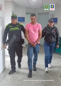 En la fotografía se aprecia en acción de caminata por un pasillo a Kevin Junior Carmona Arango. Lleva los brazos adelante y esposado. Viste camiseta de manga corta rosada, jean azul y zapatos negros. Al lado derecho de él hay un uniformado de la Policía Nacional y al lado izquierdo una servidora del CTI,