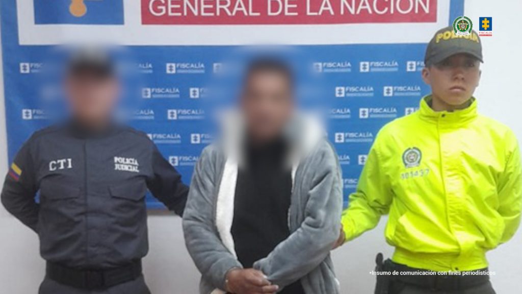 En la fotografía aparece de pie el hoy imputado. Viste con chaqueta de color gris con blanco, camiseta de color negro; jean azul oscuro y zapatillas blancas con negro. Al lado derecho de él hay un servidor del CTI de la Fiscalía y al lado izquierdo hay un uniformado de la Policía Nacional. Detrás de ellos hay un pendón institucional de la Fiscalía.