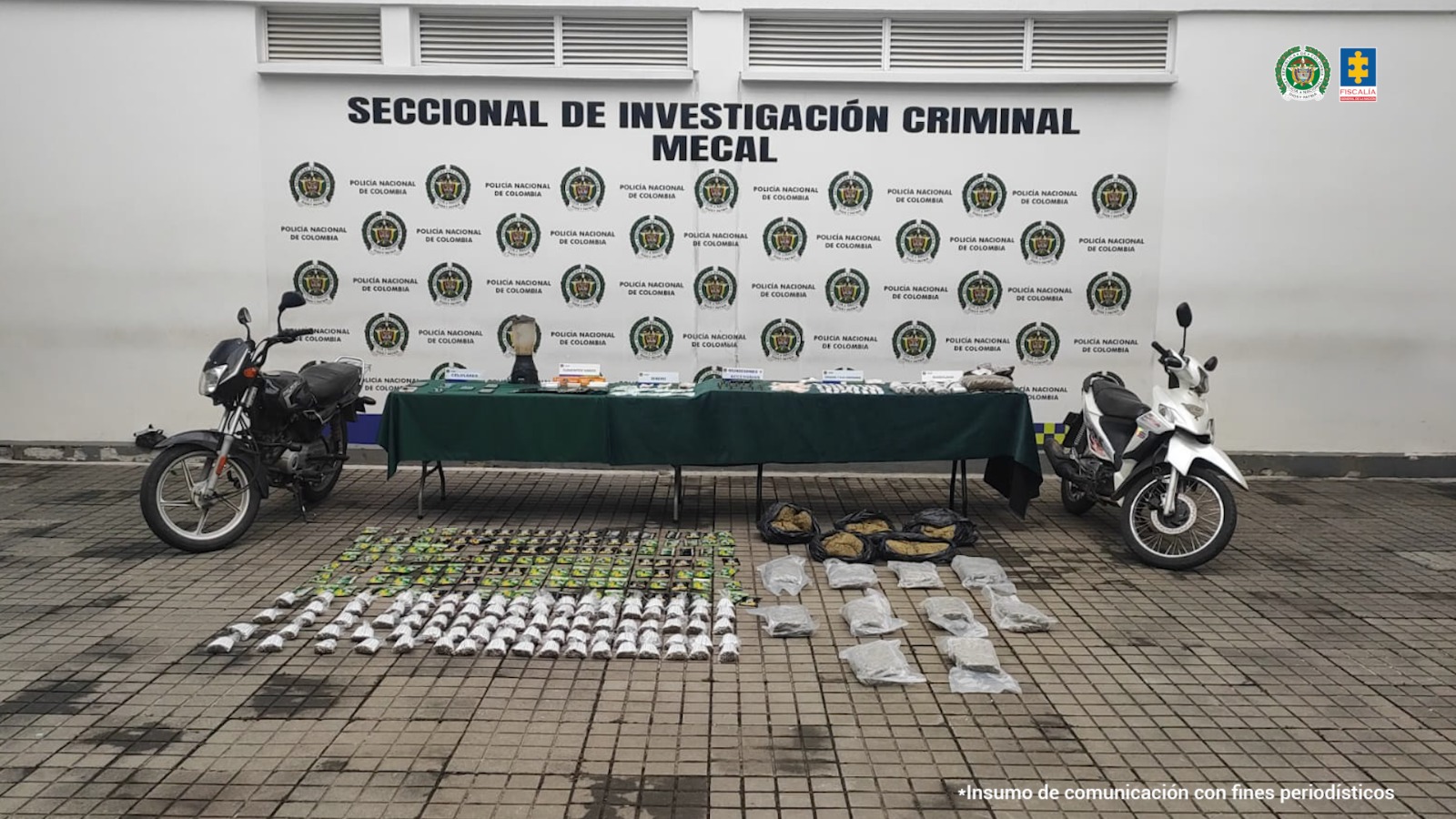 En la fotografía se aprecia una mesa con mantel verde y sobre ella gran cantidad de elementos como celulares, una licuadora, bolsas plásticas con contenido blanco y verdoso. A los extremos de la mesa hay dos motocicletas. En el suelo se aprecian varios paquetes que contienen cigarrillos y una sustancia de color café, así como marquillas de color verde con negro.
