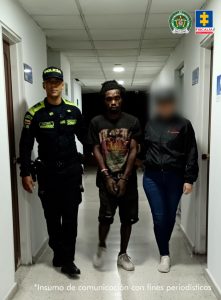En la fotografía se aprecia en acción de caminata, en un pasillo, con los brazos adelante y esposado al judicializado. Viste camiseta de manga corta de color negro con estampado de colores; bermuda negra y zapatillas blancas. Al lado izquierdo de él hay una servidora del CTI de la Fiscalía; al lado derecho de él un uniformado de la Policía Nacional.