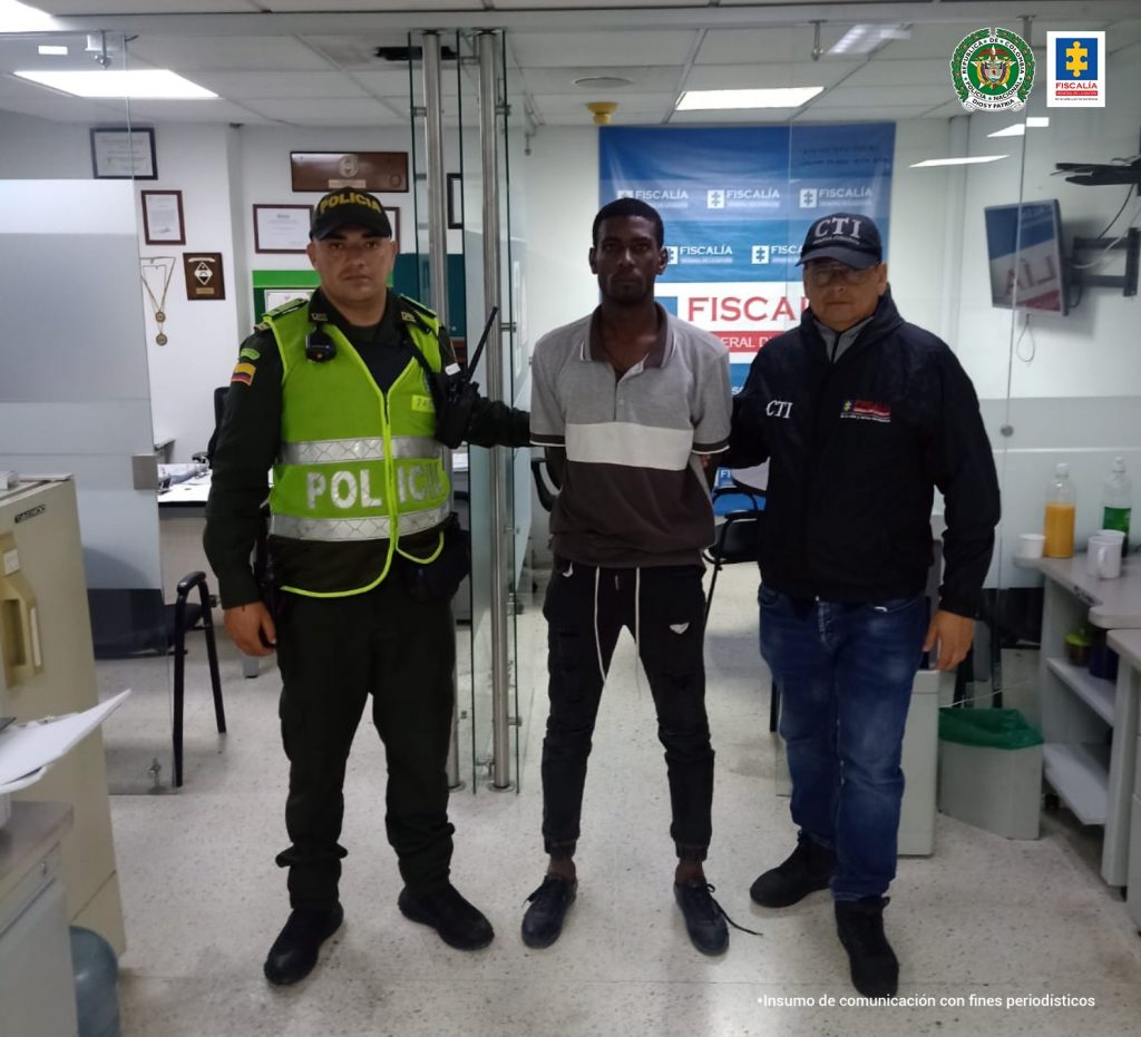 En la foto aparece, de pie y con los brazos atrás, Merlín Antonio Ramírez Urrutia. Viste camiseta de manga corta gris y negra, con franja horizontal blanca en el centro; pantalón y zapatos oscuros. Al lado derecho de él hay un uniformado de la Policía Nacional y al lado izquierdo un servidor del CTI de la Fiscalía. Detrás de ellos se aprecia un pendón de la Fiscalía.