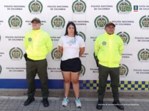 Cárcel para un hombre y una mujer como presuntos responsables del homicidio de dos adultos y la tentativa de homicidio contra dos menores de edad en Cali En la fotografía aparece de pie y con los brazos atrás Nikool Dayana Dávila Gómez. Viste camiseta blanca con estampado de una zapatilla, short negro y zapatillas blancas con cordones azules. A lado y lado de ellos hay uniformados de la Policía Nacional. Detrás de ellos hay un pendón institucional de la Policía.