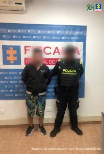 El capturado se encuentra de frente y custodiado por un uniformado de la Policía Nacional. Detrás de la foto hay un backing de la Fiscalía y el capturado tiene una chaqueta negra, bermudas y tennis negros.