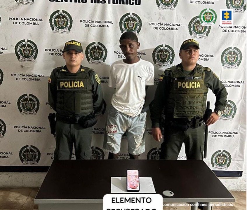 En la fotografía está un hombre con camiseta blanca, bermuda de jean, custodiado por dos agentes de la Policía Nacional. Detrás de ellos hay un pendón de esa institución y delante de ellos una mesa donde se encuentra el celular incautado.