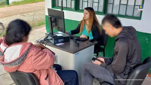 En la imagen se ve una funcionaria de la entidad de blusa verde, atendiendo a dos usuarios, una de ellas tiene una chaqueta color rosa y el otro usuario tiene una chaqueta negra y un celular en la mano.