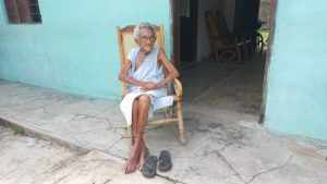 En la imagen se observa una adulta mayor sentada en una silla, frente a su casa.