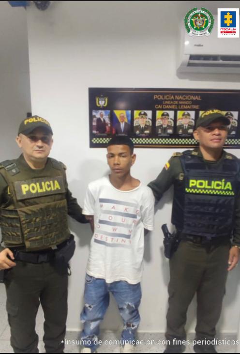 En la imagen hay un hombre joven vestido de camiseta blanca y jean azul con los brazos atrás. Se encuentra custodiado de dos agentes de la Policía Nacional.