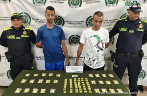 En la foto están dos hombres delgados, trigueños, de pie, de frente con los brazos esposados, cabellos negros, uno tiene camiseta azul y el otro camiseta blanca. Se encuentran con dos uniformados de la Policía Nacional y delante de ellos hay una mesa verde donde se encuentra la droga incautada.