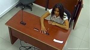  En la fotografía está el medio cuerpo de una mujer joven, trigueña de cabellos crespos negros sueltos, se encuentra sentada frente a un escritorio de madera con micrófono, vestida con un buzo blanco en la audiencia en la que se le procesó.