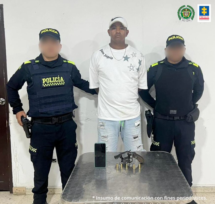 Hay un hombre moreno de pie en la fotografía con los brazos atrás, vestido de camiseta blanca, bermuda de jean y gorra blanca. Se encuentra acompañado de dos policías. Delante de ellos hay una mesa donde está el arma incautada y un celular.