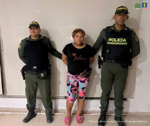 En la foto está la imagen de una mujer de contextura gruesa, trigueña, cabello café claro, vestida con una camiseta negra y una licra estampada de colores azul, negra y rosada con unas sandalias rosadas. Se encuentra custodiada por dos agentes de la Policía Nacional.