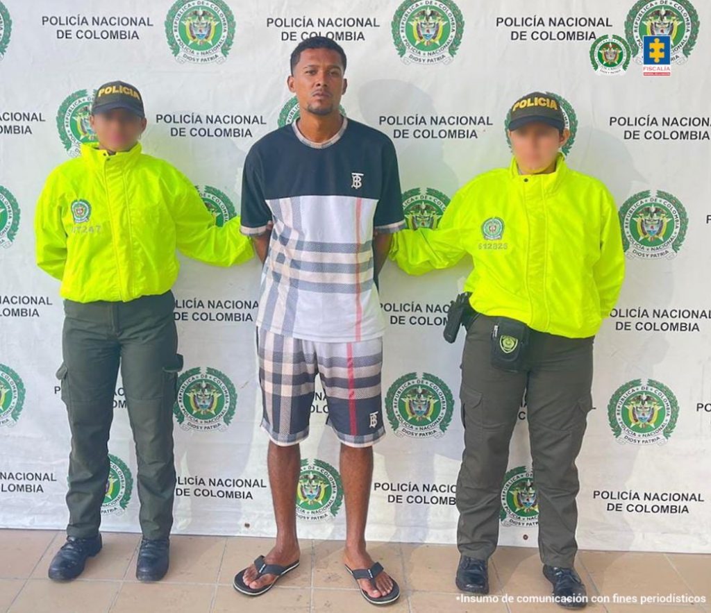 En la foto está un hombre alto, moreno, de pie, con los brazos atrás, vestido de camiseta y bermuda azul de cuadros, sandalias negras, acompañado por dos uniformados de la Policía Nacional. Detrás de ellos hay un pendón de esa institución.