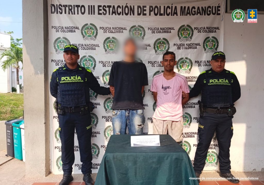 En la foto aparecen dos hombres de pie, delgados con los brazos atrás esposados, uno es alto y está vestido con camiseta negra y jean azul y el otro es de baja estatura vestido con una camiseta rosada y un pantalón beige. Estan custodiados por uniformados de la Policía Nacional.
