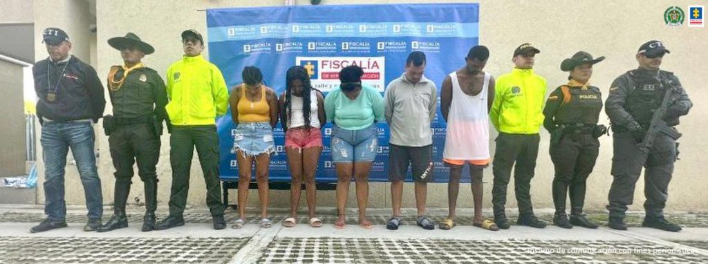 En la imagen se encuentran tres mujeres y dos hombres de pie con las cabezas hacia abajo y los brazos atrás. Las personas se encuentran custodiadas por uniformados del CTI de la Fiscalía y la Policía Nacional. Detrás de ellos hay un pendón de la Fiscalía General de la Nación.