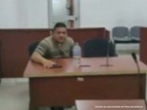 En la foto aparece un hombre sentado frente a un escritorio de madera con micrófono, vestido de camiseta marrón y rallas verdes en el desarrollo de la audiencia donde se le procesó.