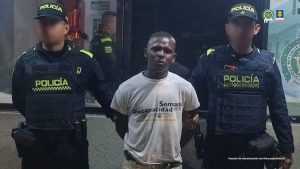 En la fotografía está un moreno, de pie, con los brazos atrás, de contextura gruesa, de color, con los cabellos cortos crespos negros, vestido de camiseta blanca y pantalón beige, custodiado por dos uniformados de la Policía Nacional.